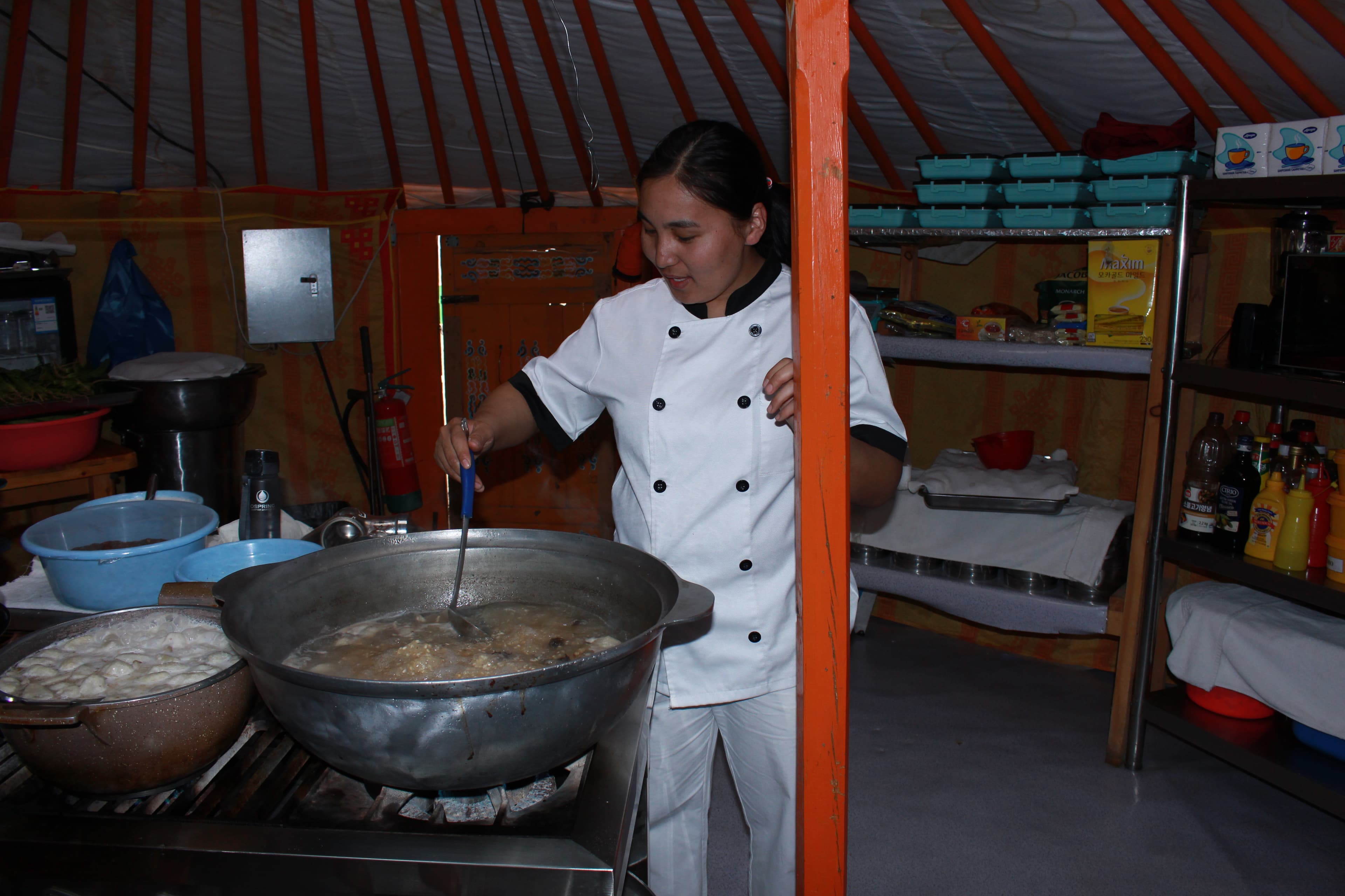 A cook in a white uniform stirs a large metal pot in a kitchen with exposed wooden supports